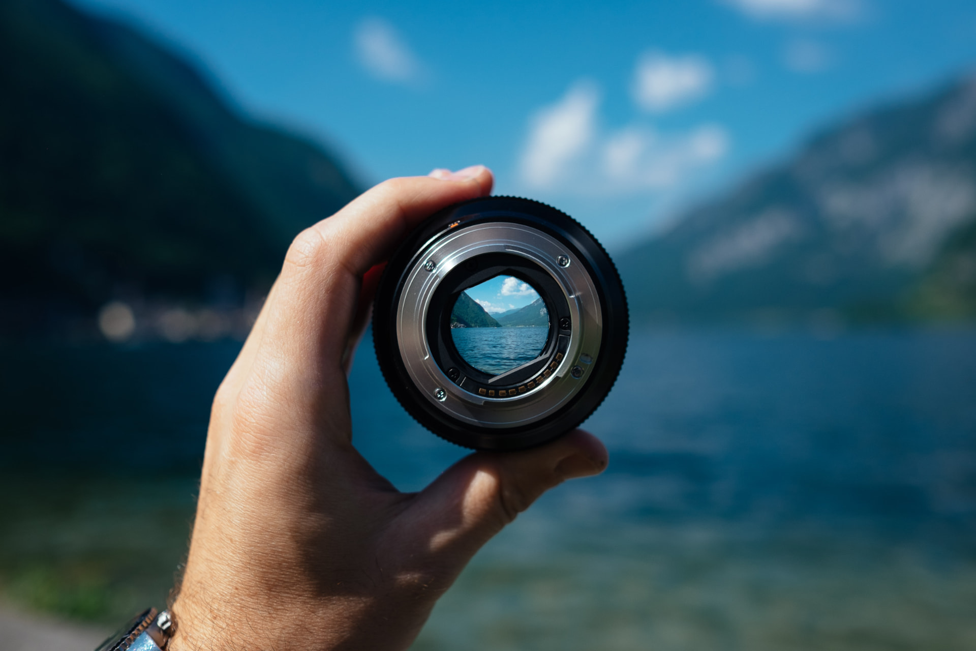 A hand holding a camera lens that brings a mountain lake into sharp focus
