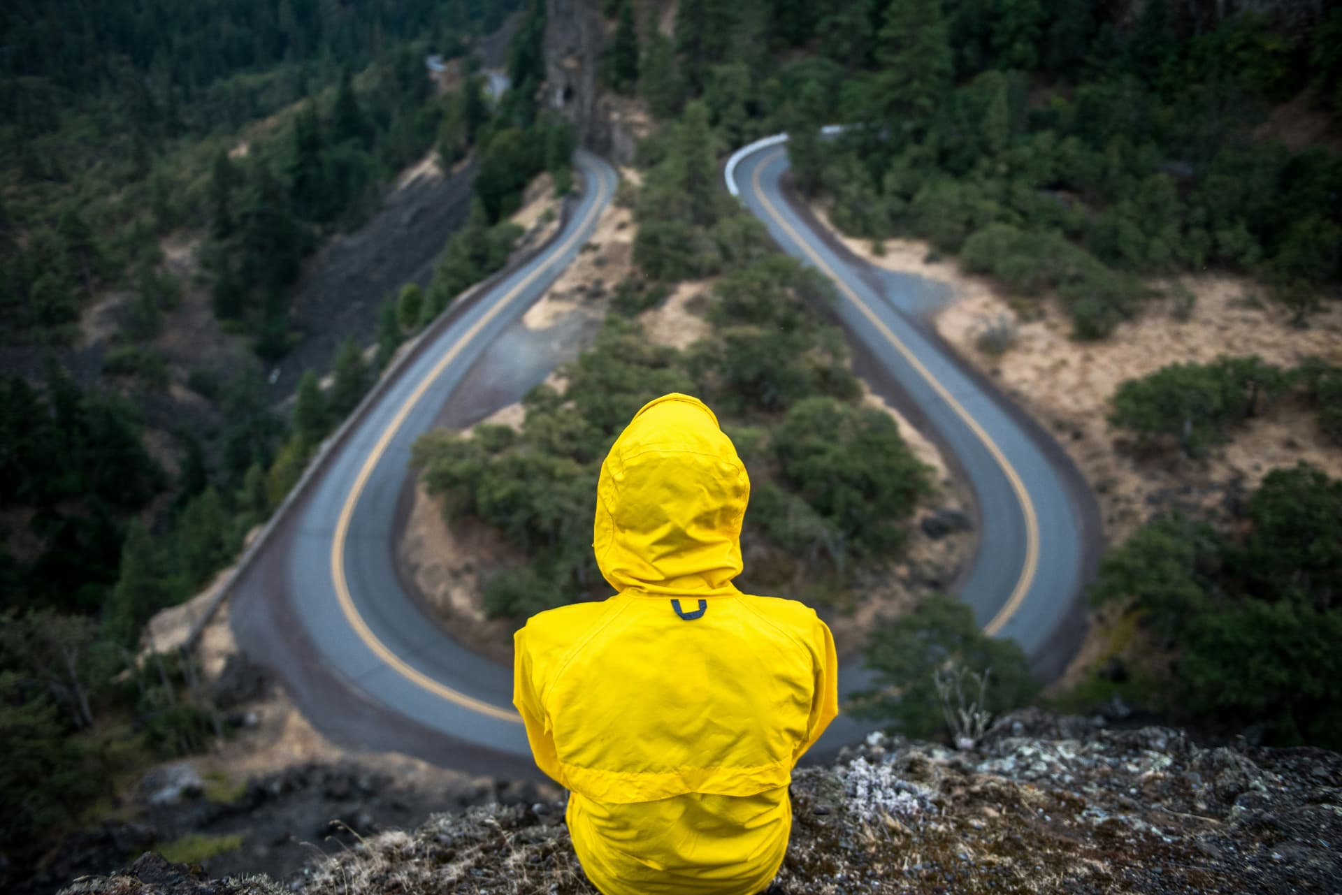 A person in a yellow jacket sitting on a cliff edge above a horseshoe bend in a mountain road