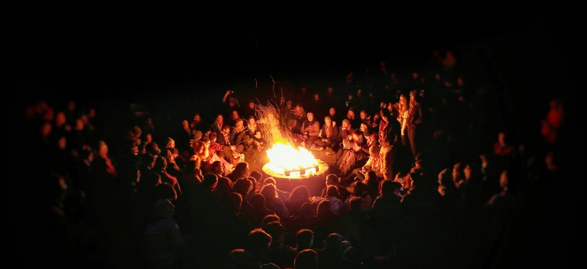 A crowd of people seated in a circle around a bonfire at night